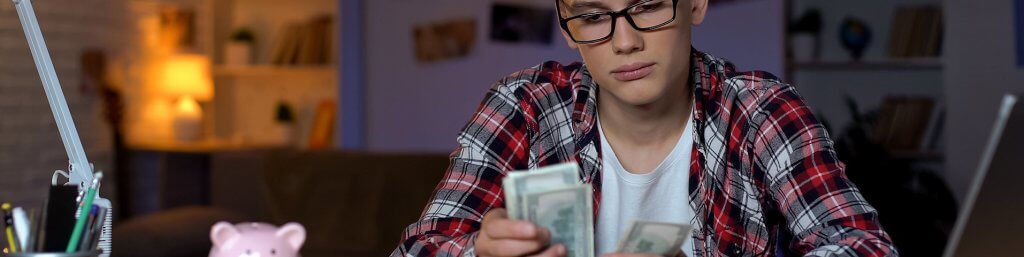 Teenage boy sitting at a desk counting several dollar bills with a piggy bank next to him